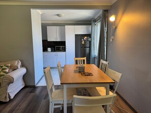 A dining area with a wooden table and white chairs, a kitchen in the background with white cabinets and appliances, and a grey wall with a light fixture. - Entire villa in Metford, 
A relaxing, homey stay — ideal for short breaks (Metford)
