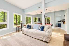 A spacious living room with a large sofa, multiple windows, a pool table, and a view of the outdoors. - 🌊 Beechnut Cove | Lake Gaston Escape • Private Dock • Game Room • Fire Pit 🔥 (Littleton)