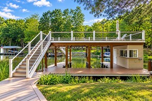 A wooden deck with a staircase leading to a covered area with a railing, overlooking a calm body of water with boats. - 🌊 Beechnut Cove | Lake Gaston Escape • Private Dock • Game Room • Fire Pit 🔥 (Littleton)