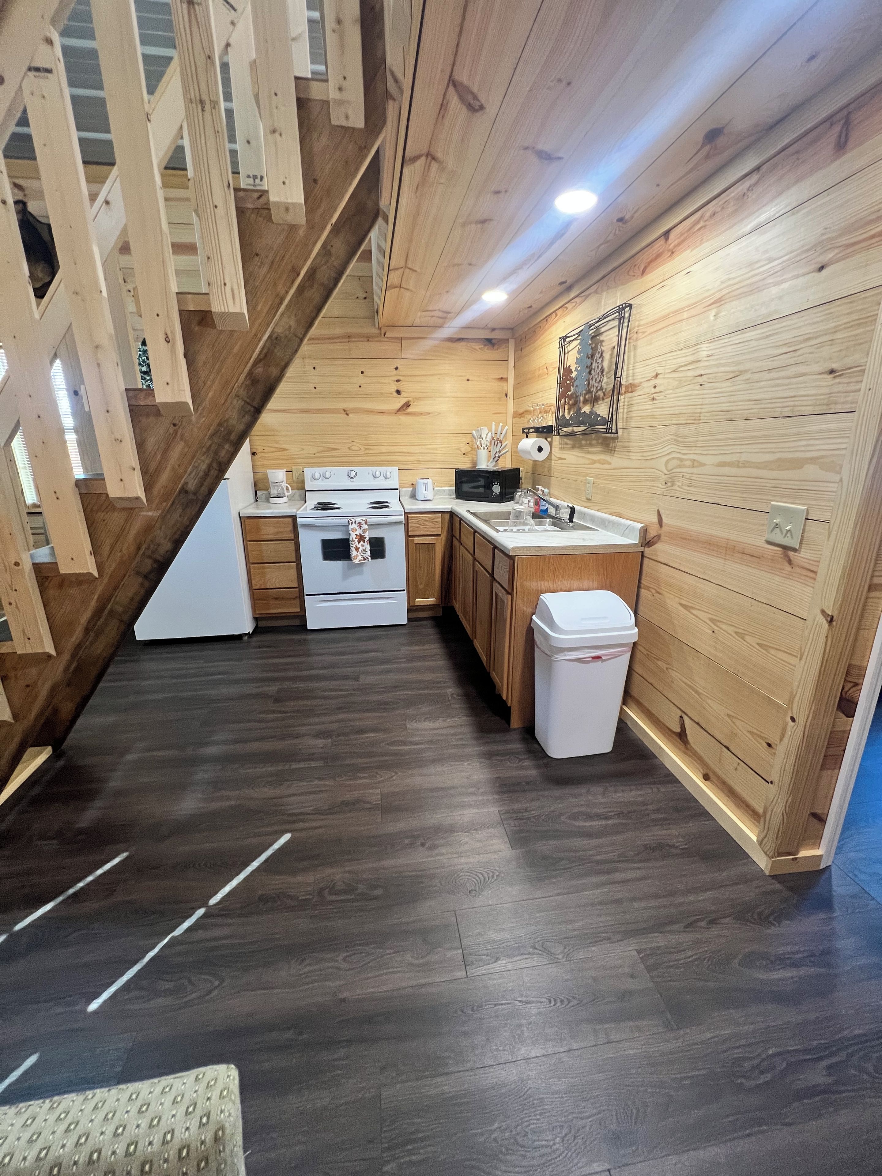 A kitchen with wooden walls, a dark wooden floor, a white stove, a sink, and a trash bin.