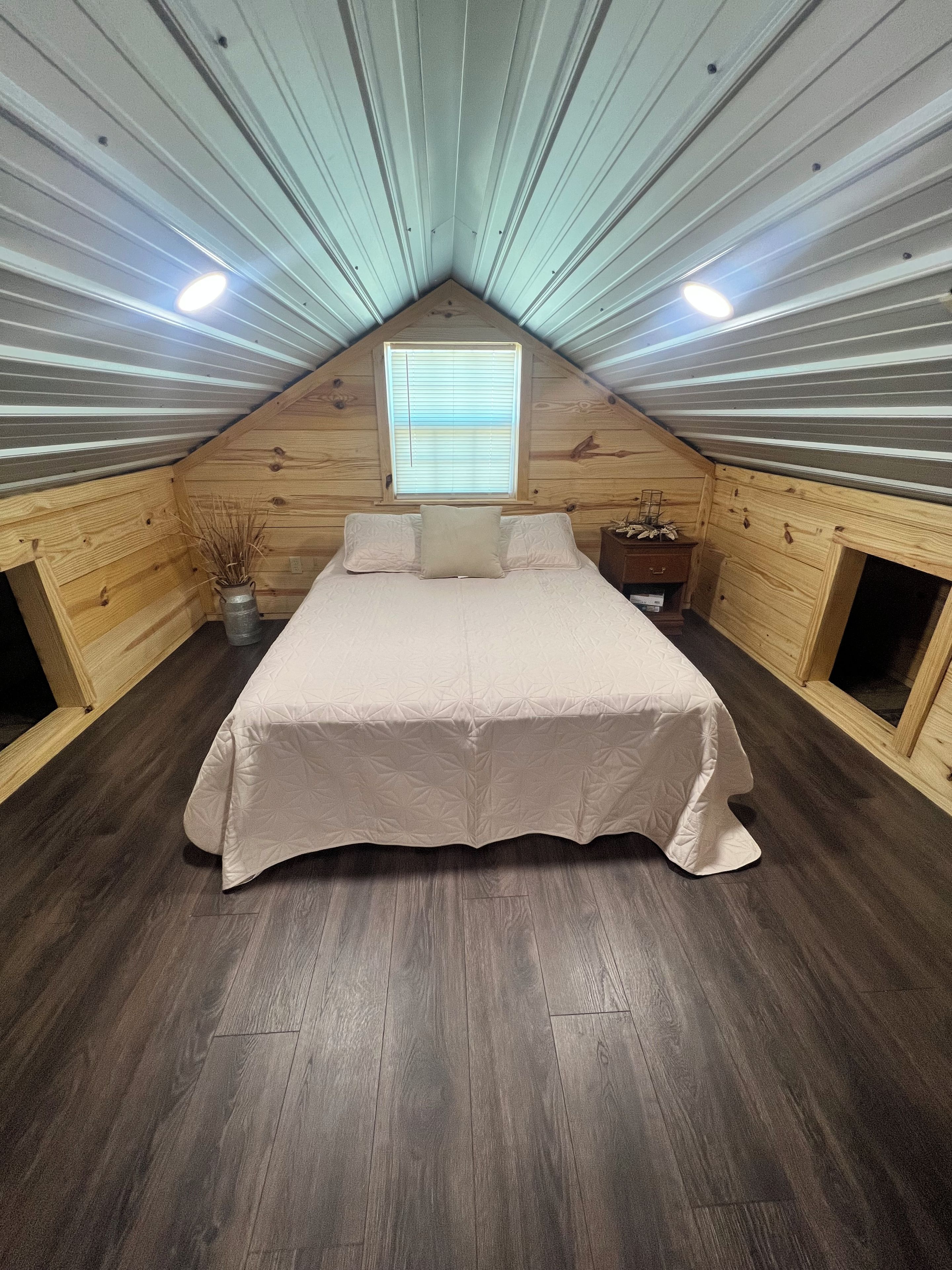 Un dormitorio con techo inclinado, una cama con cubrecama color beige, una ventana con persianas y paredes de madera.