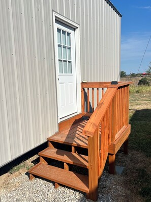 Une terrasse en bois mène à une porte blanche d’un bâtiment en métal ondulé.