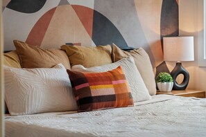 A bed with multiple pillows, a bedside table with a lamp, and a wall with abstract art. - Leave the car parked. Edmonton's best cafés & restaurants are minutes' walk away, downtown 5 by car. Your brand-new upper home: gallery walls, chef-style kitchen with a gas range, fireplace, A/C & patio. For professionals & families, room for 9. (Edmonton)