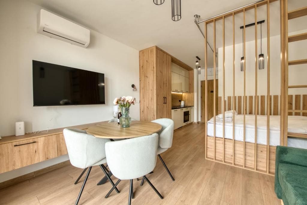 A modern room with a wooden dining table, white chairs, a flat-screen TV mounted on the wall, a kitchen area, and a bed with a wooden headboard.