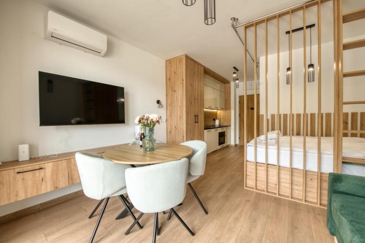 Een moderne kamer met een houten eettafel, witte stoelen, een flatscreen televisie aan de muur, een keukenhoek en een bed met een houten hoofdeinde.