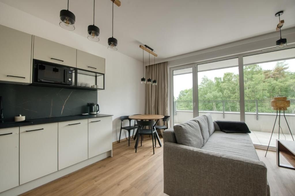 A modern kitchen with a microwave, dining table, and chairs, a sofa, and large windows with a view of greenery.