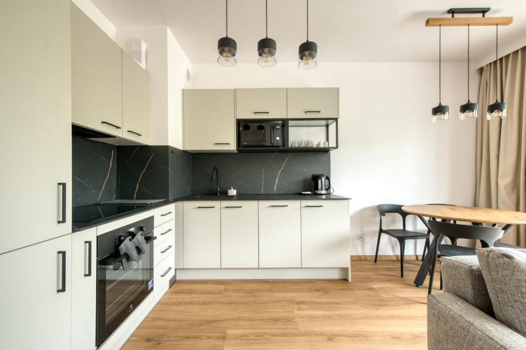 A modern kitchen with white cabinets, a black countertop, and a built-in oven. There is a dining area with a wooden table and chairs.