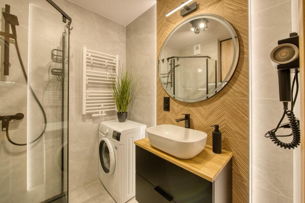 A modern bathroom with a glass shower enclosure, a built-in washing machine, a wooden vanity with a vessel sink, and a round mirror.