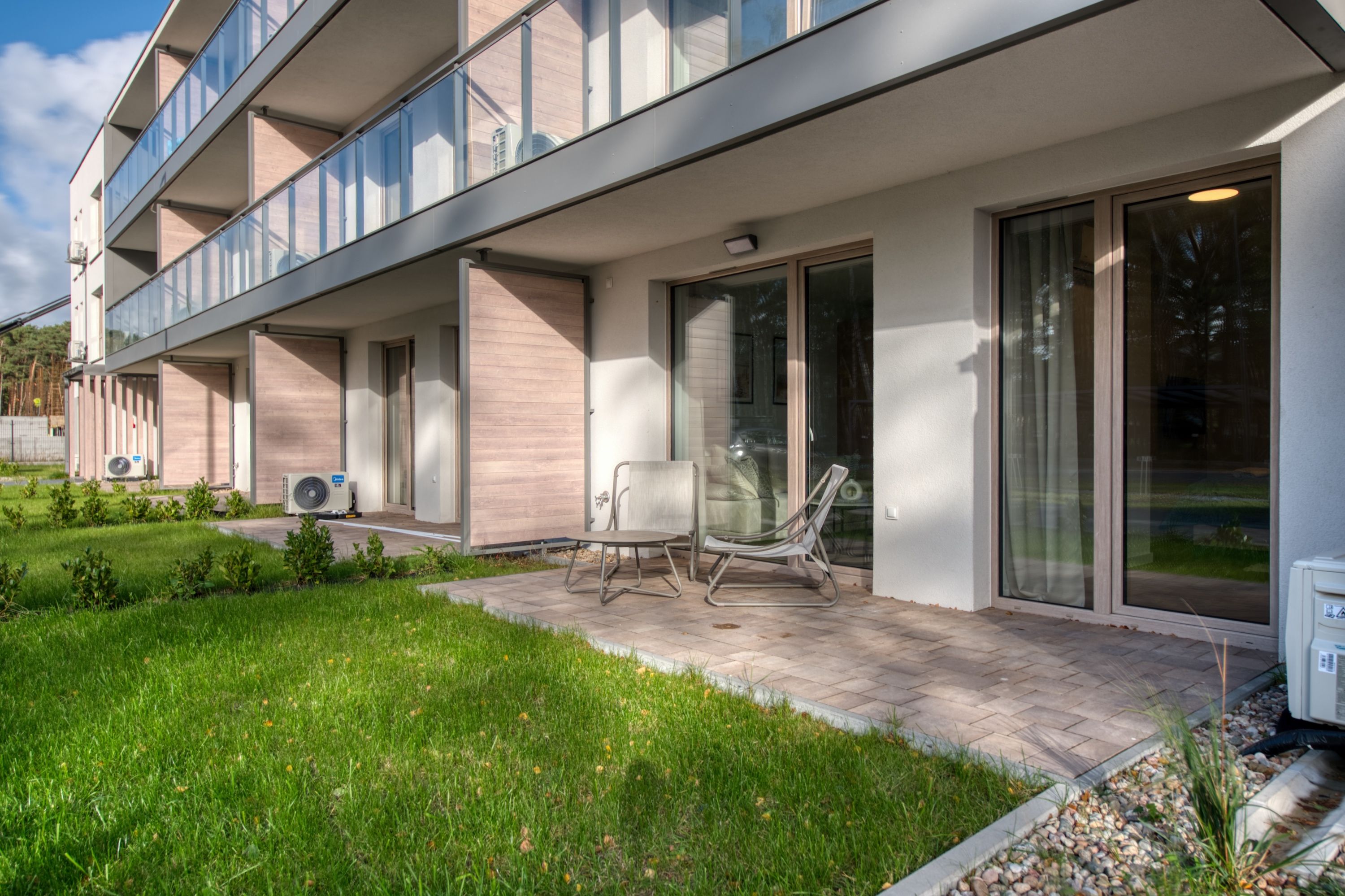 Ein modernes Gebäude mit flachem Dach, großen Fenstern und einer gepflasterten Terrasse mit Gartenmöbeln.