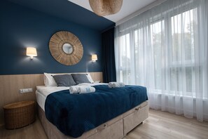 Una habitación de hotel moderna con una cama grande, un espejo redondo, dos lámparas de pared y un amplio ventanal con cortinas translúcidas.