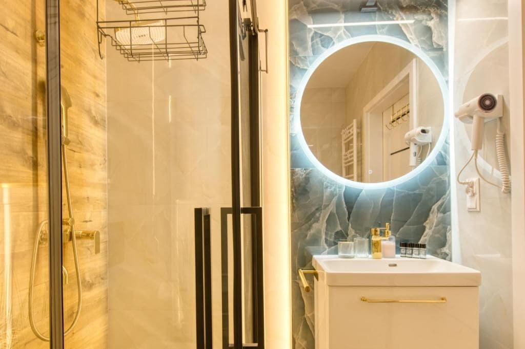 A bathroom with a glass-enclosed shower, a vanity with a marble countertop, and a round mirror with a lighted frame.