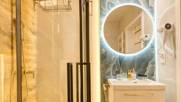A bathroom with a glass-enclosed shower, a vanity with a marble countertop, and a round mirror with a lighted frame.
