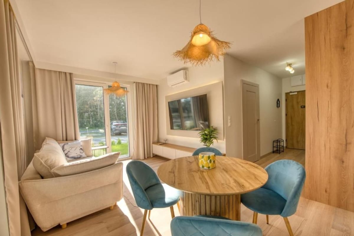 A modern living room with a round wooden table, blue chairs, a sofa, and a large window with curtains.
