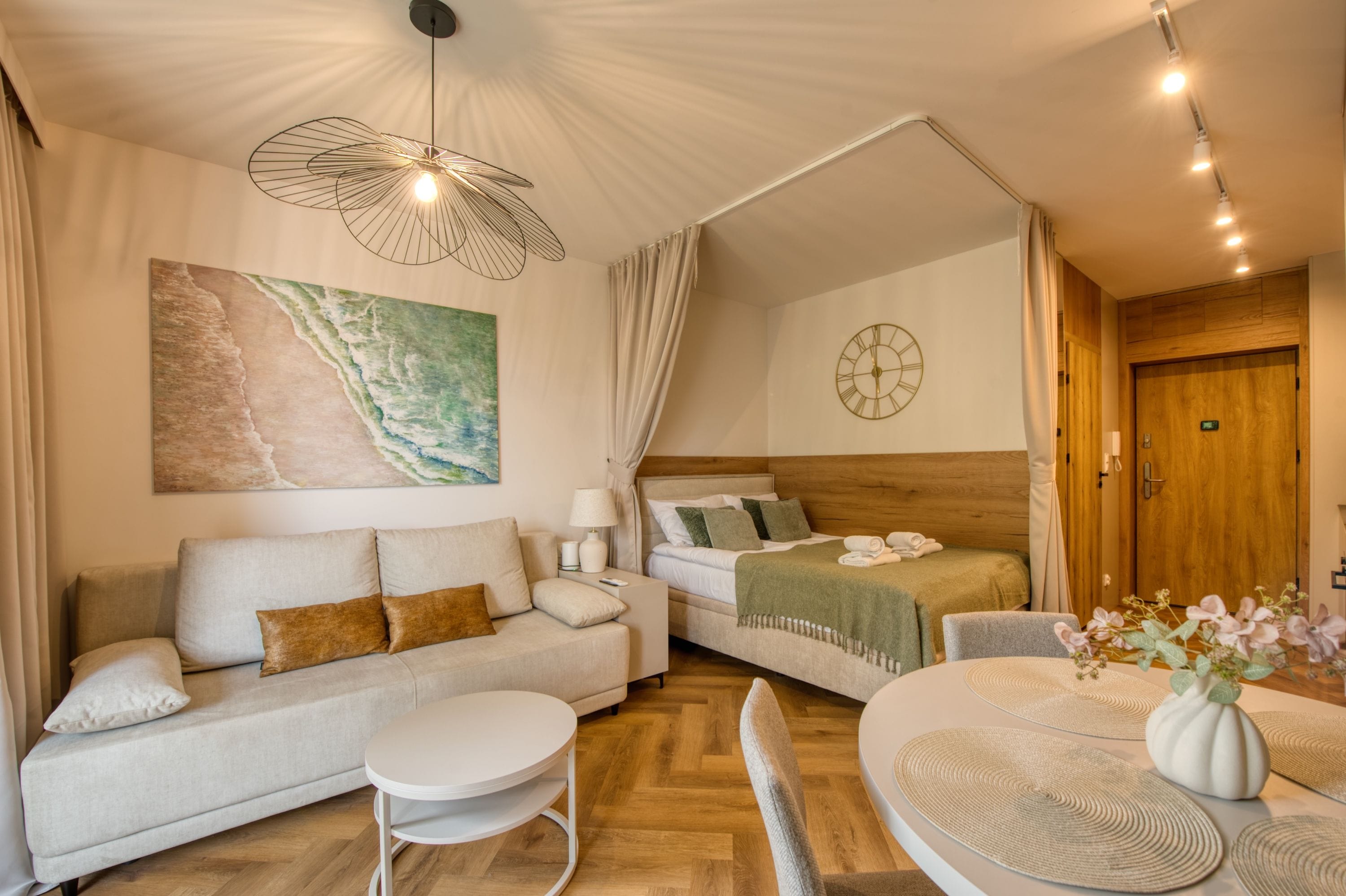 A room with a bed, a sofa, a small table, a wall clock, and a painting.