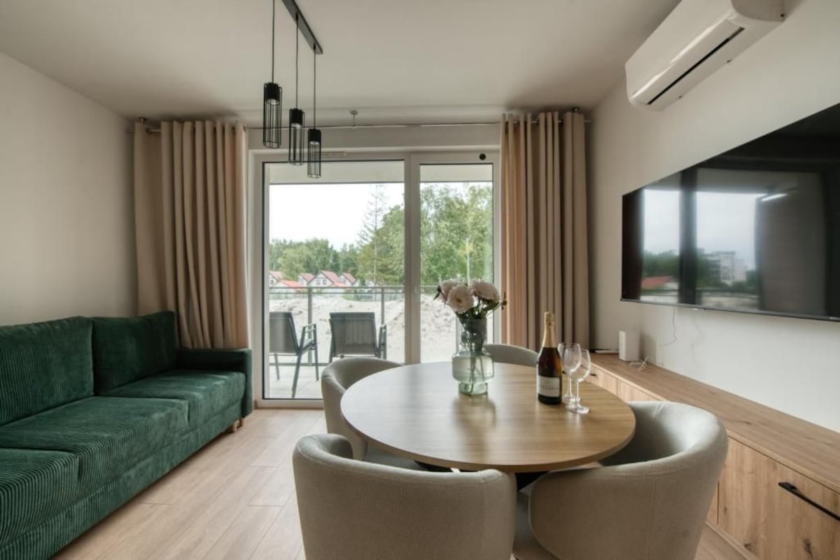 A modern living room with a green sofa, a round wooden table with chairs, a vase of flowers, and a flat-screen TV mounted on the wall.