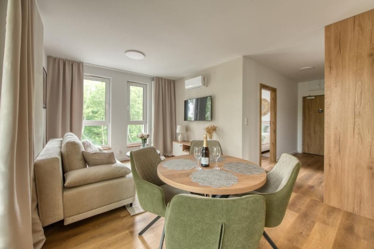 A modern living room with a sofa, a dining table with chairs, a TV mounted on the wall, and a wooden floor.