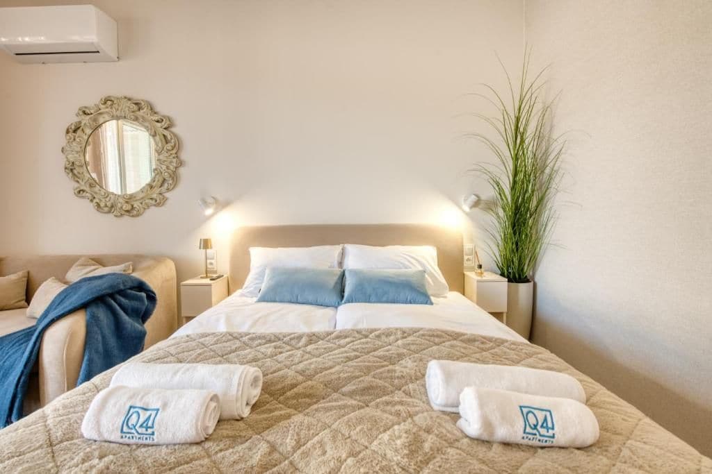 Een hotelkamer met een bed, nachtkastje, spiegel, een potplant en een handdoek met een logo.