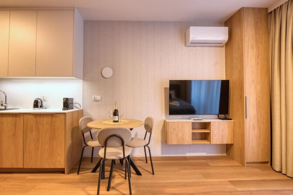A compact living space with a kitchenette, a dining area with a table and chairs, a flat-screen TV mounted on the wall, and a window with curtains.