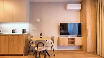 A compact living space with a kitchenette, a dining area with a table and chairs, a flat-screen TV mounted on the wall, and a window with curtains.