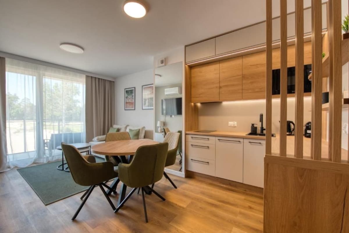 Een moderne keuken met houten kasten, een eethoek met een ronde tafel en stoelen, en een groot raam met gordijnen.