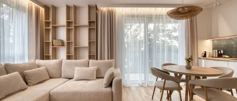 A modern living room with a sectional sofa, a dining table with chairs, a built-in bookshelf, and a large window with sheer curtains.