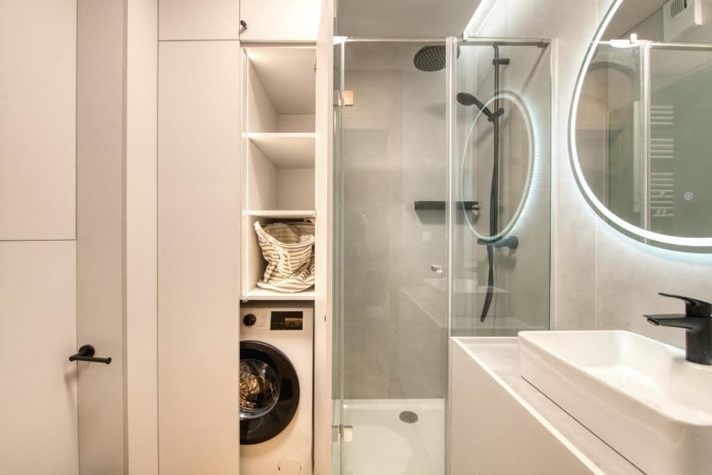 A modern bathroom with a glass-enclosed shower, a built-in washing machine, and a white sink with a mirror.