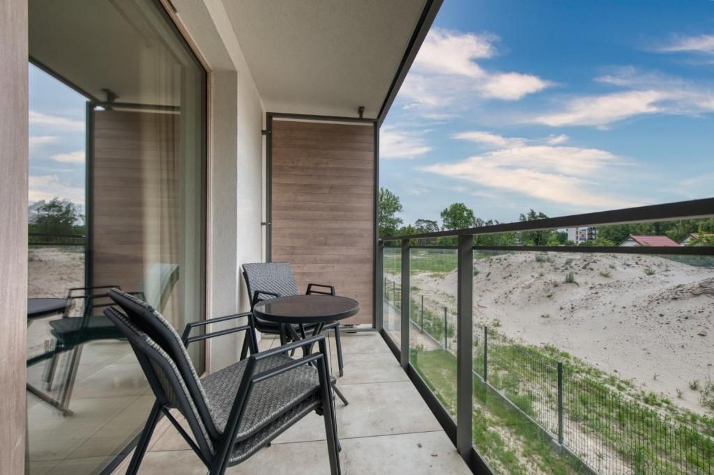 A balcony with a table and chairs, a glass door, and a view of a sandy area with a fence.