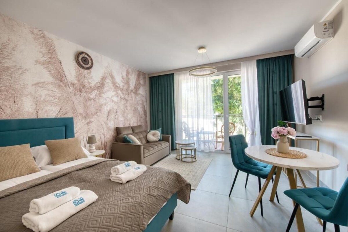 Habitación de hotel con cama, sofá, zona de comedor con sillas, televisor y balcón con cortinas.
