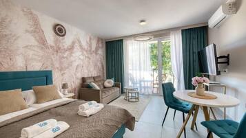 A hotel room with a bed, a sofa, a dining area with chairs, a TV, and a balcony with curtains.