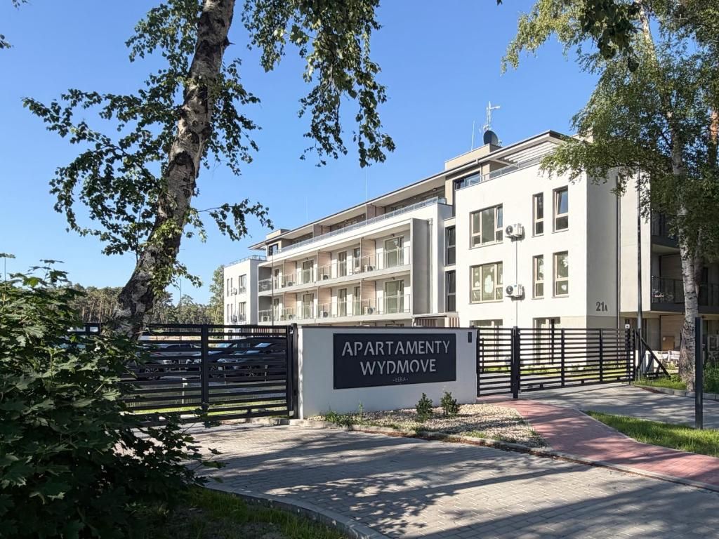 A multi-story apartment building with a gated entrance, a sign indicating 'APARTMENTY WYDMOVE,' and a paved pathway leading to the entrance.