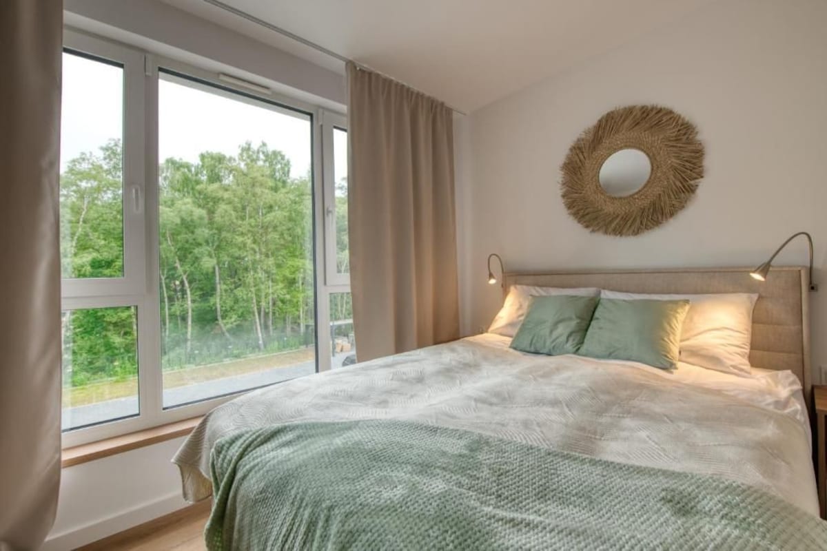 Een slaapkamer met een groot raam, een bed met een groene deken, een ronde wanddecoratie en een bedlampje.