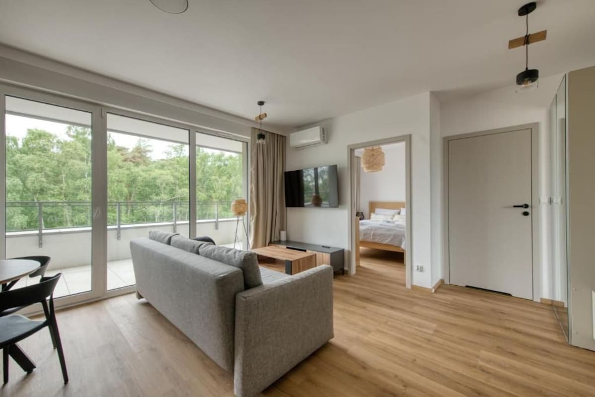 A modern living room with a sofa, a dining table, and a TV. There is a balcony with large windows and a door leading to another room.