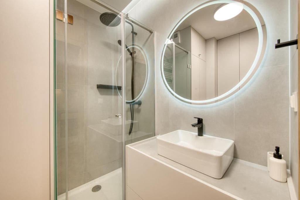 Ein modernes Badezimmer mit einer gläsernen Dusche, einem weißen Waschbecken und einem runden Spiegel mit beleuchtetem Rand.