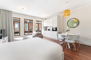 Ein modernes Hotelzimmer mit einem großen Bett, einem Essbereich mit Tisch und Stühlen, einer Küche und großen Fenstern mit Vorhängen.