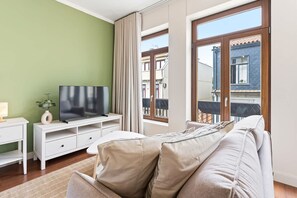 Ein modernes Wohnzimmer mit einem Flachbildfernseher auf einem weißen TV-Möbel, einem beigen Sofa und einem großen Fenster mit Blick auf Gebäude.