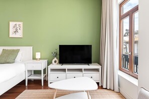 Ein Schlafzimmer mit Bett, Nachttisch, Fernseher auf einem Schrank und einem Fenster mit Vorhängen, Blick auf Gebäude im Freien.
