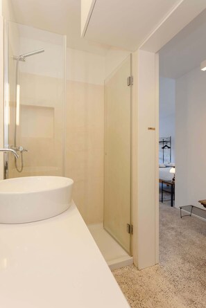 A modern bathroom with a white sink, glass shower enclosure, and a visible bedroom with a bed and a nightstand. - Liiiving in Porto - Lobo Studio I (Porto)