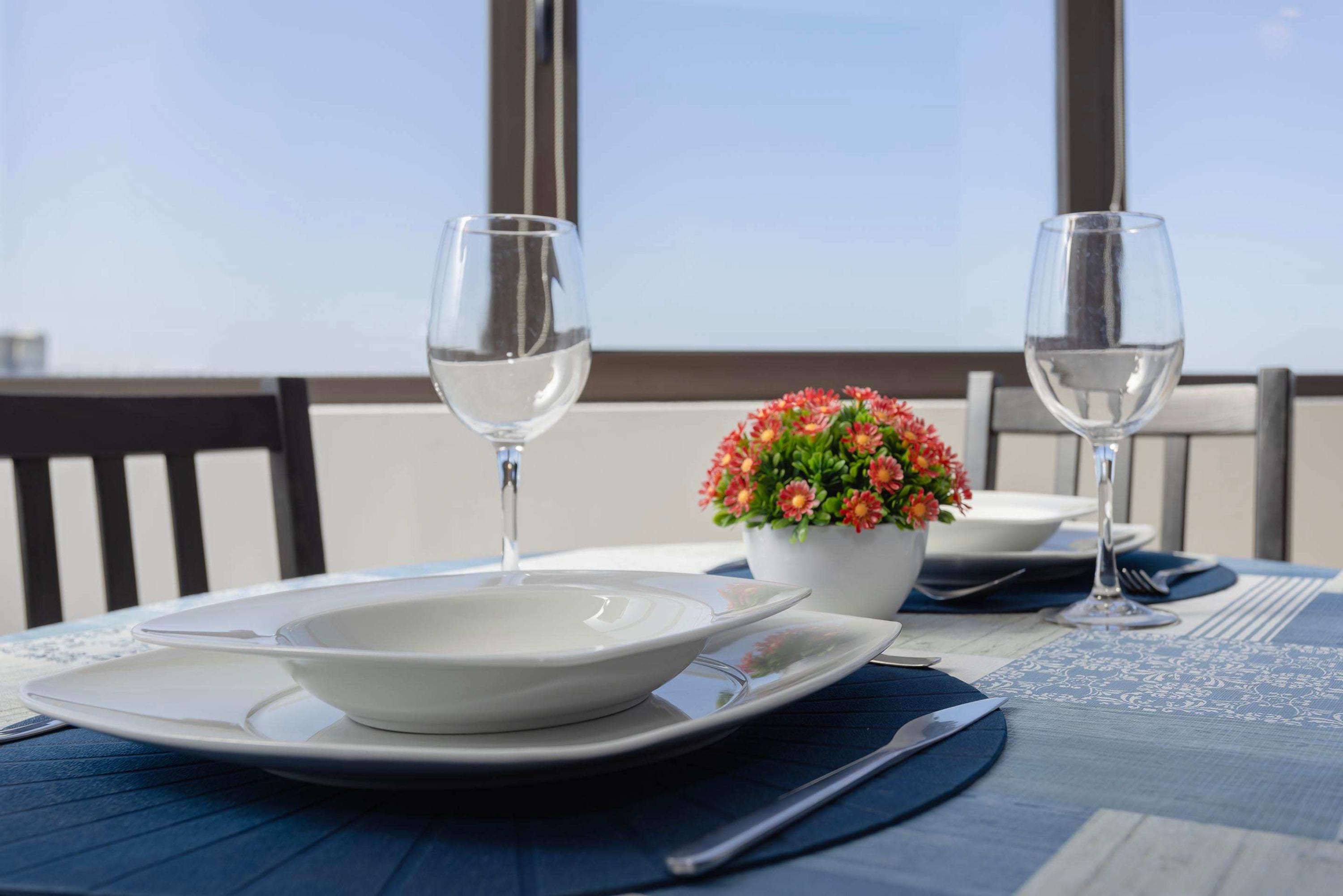 A table set with a white plate, a bowl, two wine glasses, a knife, and a fork on a blue tablecloth.