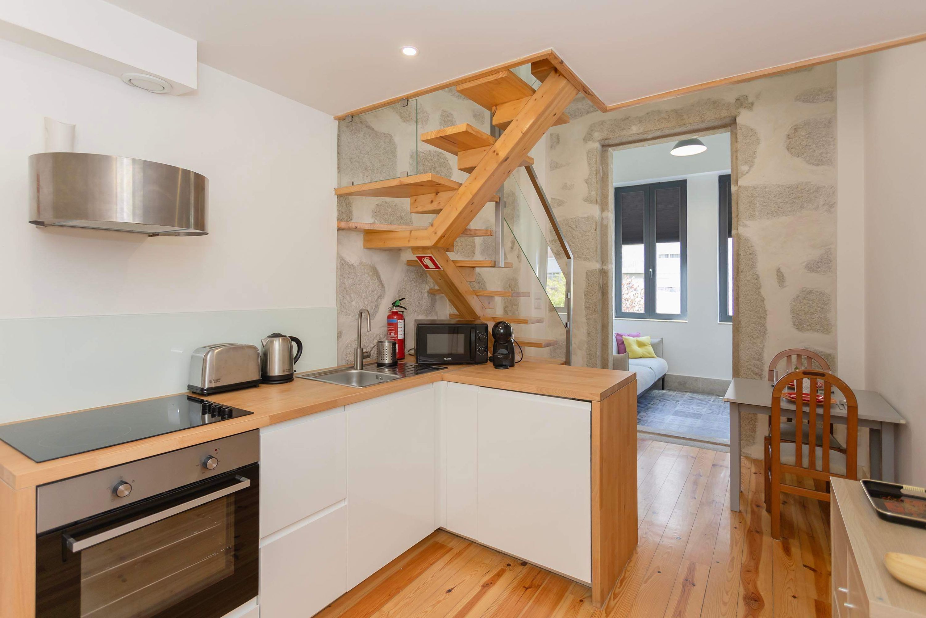 A compact kitchen with a built-in oven, a microwave, and a toaster. A wooden staircase leads to an adjacent living area.