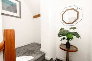 A staircase with a wooden handrail, a small round table with a potted plant, and a wall-mounted mirror. - Tradewinds - 2 Bedroom Apartment - Tenby (Tenby)