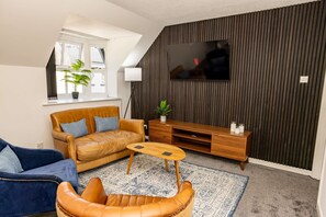 A modern living room with a sofa, armchair, coffee table, TV, and a plant. - Tradewinds - 2 Bedroom Apartment - Tenby (Tenby)