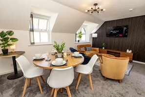 A modern living room with a dining area, a TV mounted on the wall, and a ceiling fan. - Tradewinds - 2 Bedroom Apartment - Tenby (Tenby)