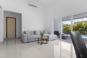 A modern living room with a grey sofa, a coffee table, and a view of a pool through a sliding glass door. - Modern 3BR Villa With Private Pool in Oasis del Lago by Vistacana Rentals (Punta Cana)