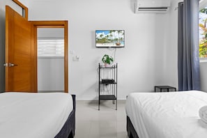 Habitación de hotel con dos camas, un televisor en la pared, una planta en un estante y una ventana con cortinas.
