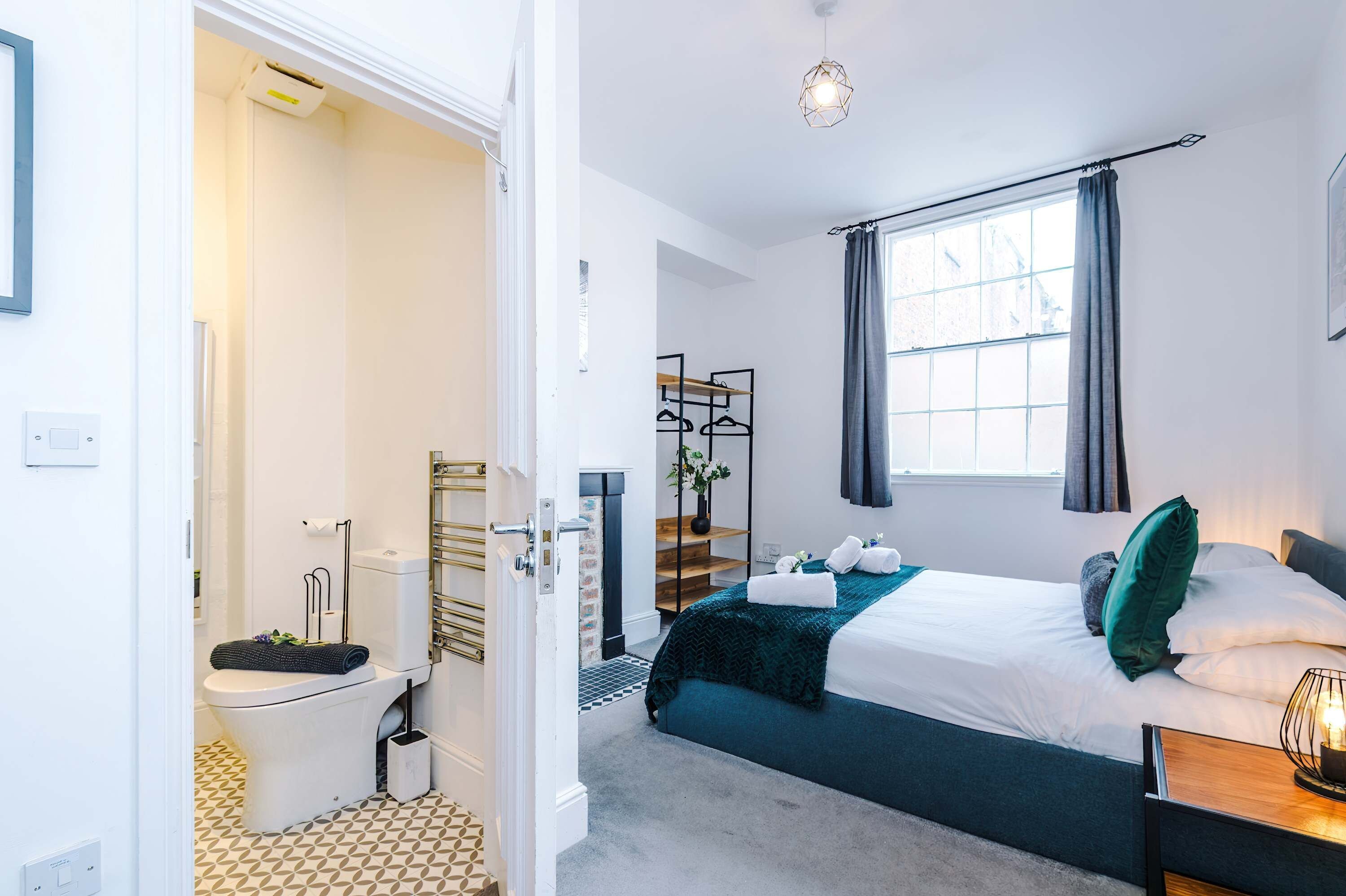 Una camera d'albergo con un letto, una finestra con tende, un comodino con una lampada, un bagno con il water e una doccia, e un corridoio con una porta.