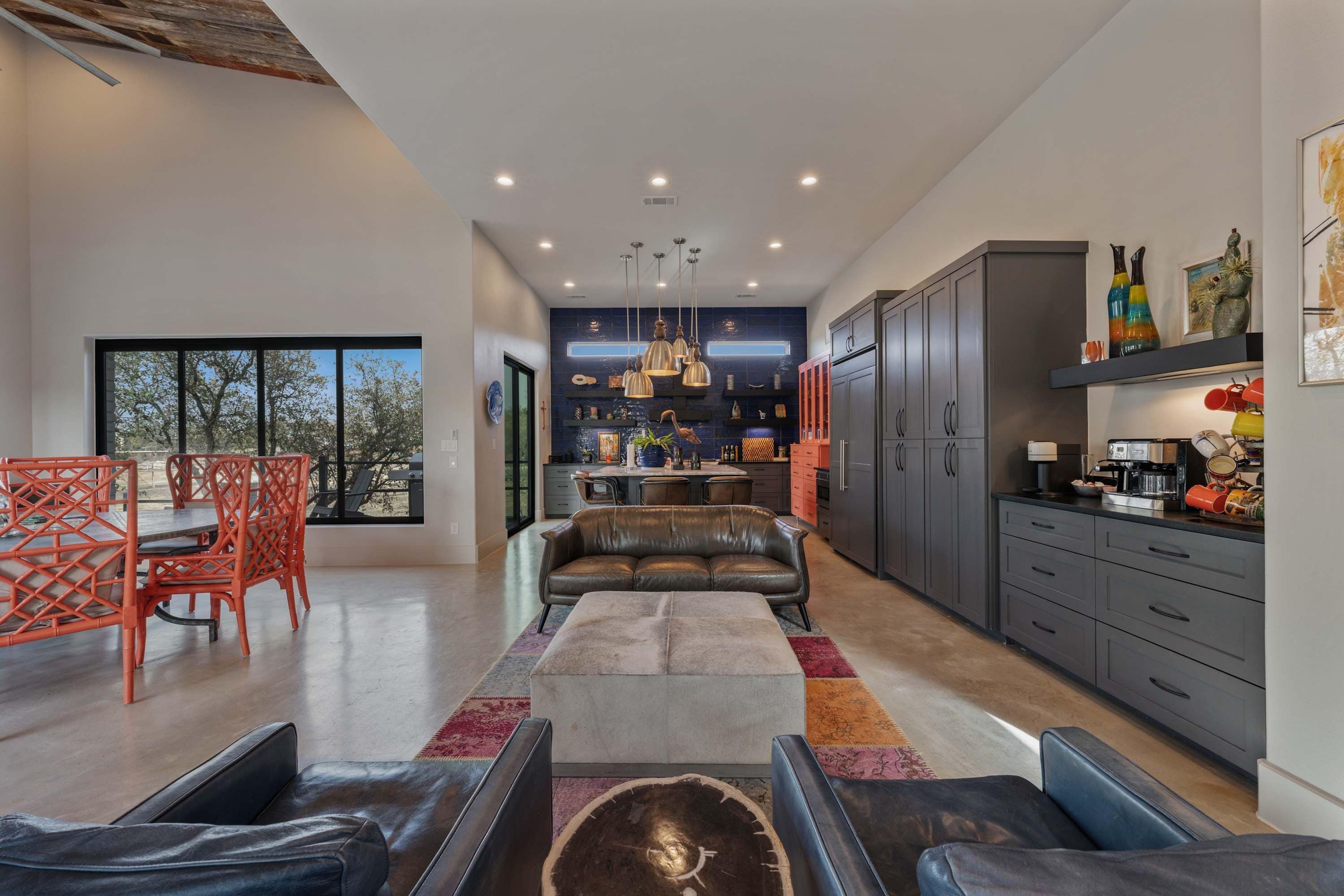 A modern kitchen with dark cabinets, a dining area with orange chairs, and a large window overlooking a tree.