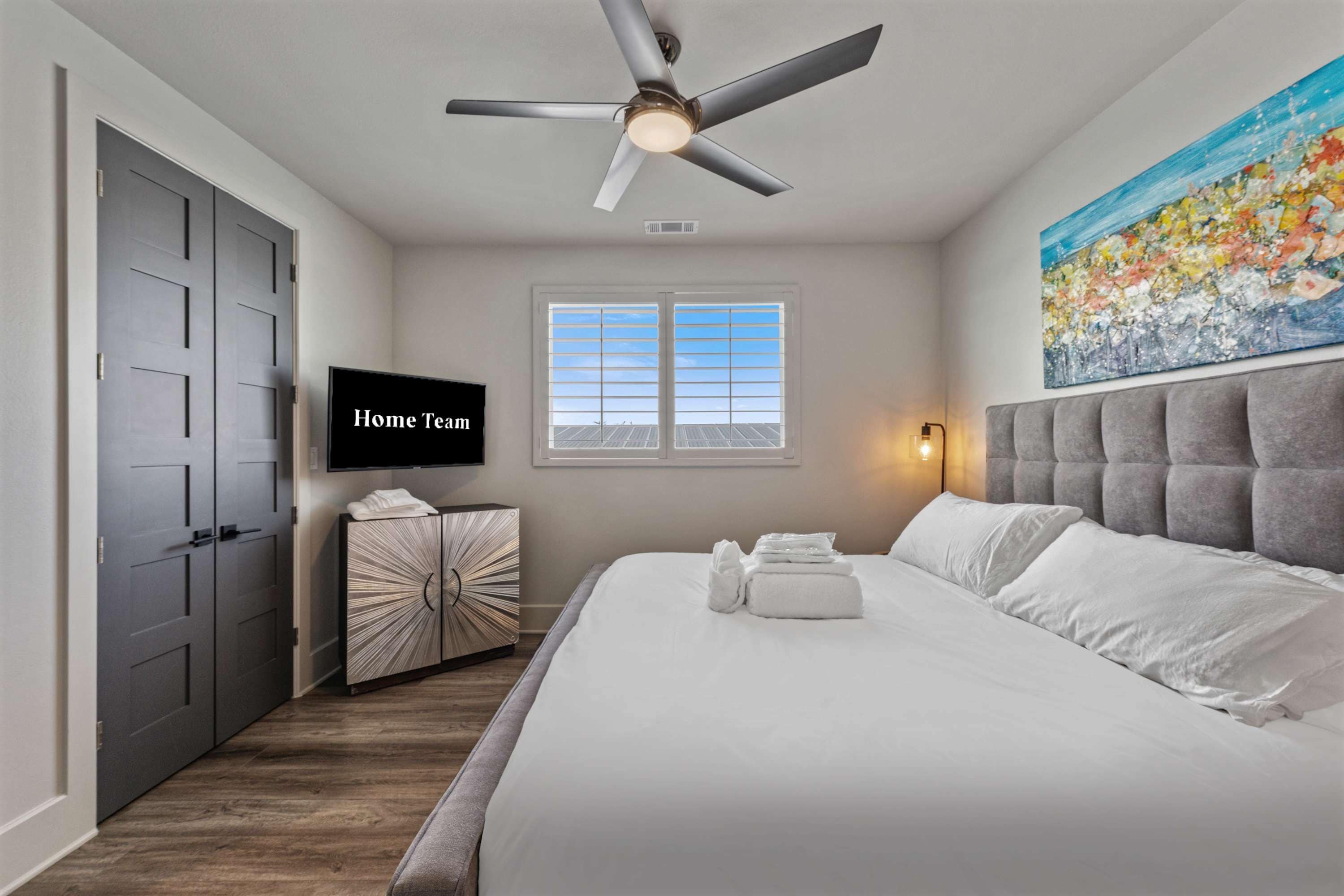 A hotel room with a bed, a television, a ceiling fan, a window, and a painting on the wall.