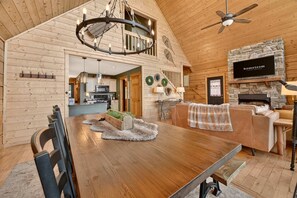 A spacious living area with a wooden dining table, a fireplace, and a television mounted on a stone wall. - Ski-out to Hatley Pointe Hot Tub Sleeps 8 (Mars Hill)