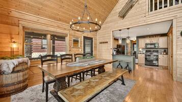 A wooden interior with a dining area featuring a large table and benches, a chandelier, and a kitchen with modern appliances.