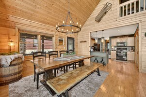 A wooden interior with a dining area featuring a large table and benches, a chandelier, and a kitchen with modern appliances.